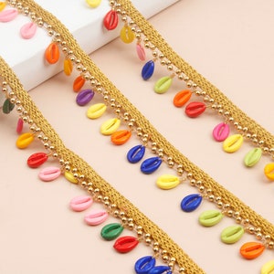 以下が含まれることがあります： 織り込まれたテクスチャの金色のトリムで、カラフルな貝殻型のビーズで飾られています。ビーズは赤、ピンク、黄色、緑、オレンジ、青など様々な色があります。左下にはハサミが見えます。