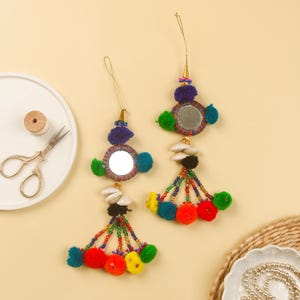 以下が含まれることがあります： 鏡、ビーズ、ポンポンが付いたカラフルなタッセルオーナメント2つ。各オーナメントには、カラフルなビーズのトリムで囲まれた丸い鏡と、上部に小さなポンポンが付いています。タッセルはカラフルなビーズとポンポンで作られています。