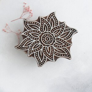 May include: A wooden block printing stamp with a detailed floral design. The design features a central circle surrounded by intricate petals and leaves. The stamp is brown with white details.