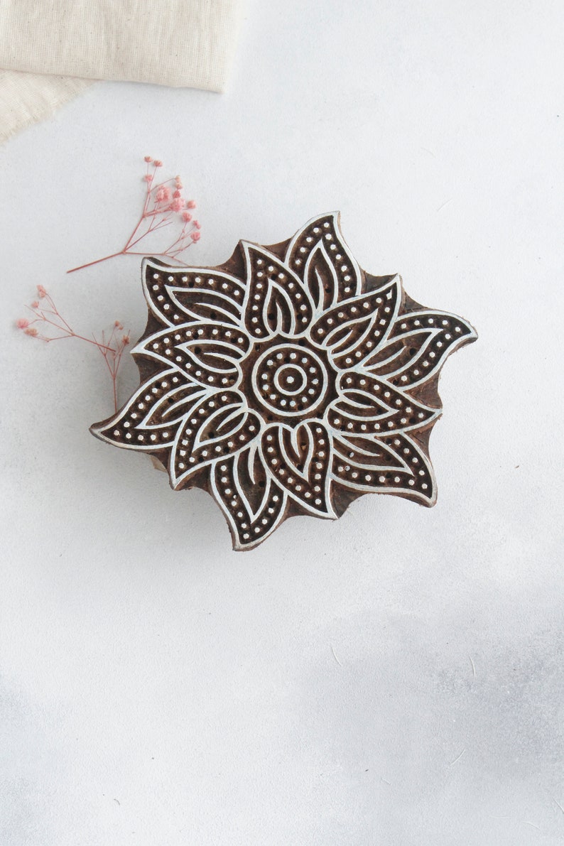 May include: A wooden block printing stamp with a detailed floral design. The design features a central circle surrounded by intricate petals and leaves. The stamp is brown with white details.