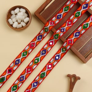 Puede incluir: Tres hebras de ribete rojo con bordados coloridos y detalles de espejo. El ribete presenta un patrón repetitivo de blanco, naranja, verde, azul y plateado.