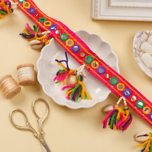 以下が含まれることがあります： ピンクのファブリックトリムにカラフルなタッセル、ミラーの装飾、白いビーズが付いています。トリムは白いプレートの上にあり、金色のハサミと糸巻きが置かれています。