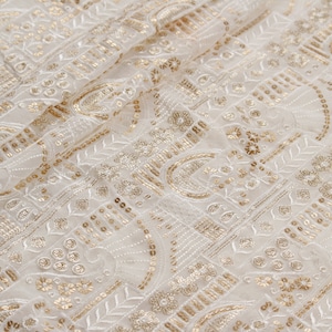 Pode incluir: Close-up de tecido marfim com bordados dourados intrincados e lantejoulas. O design apresenta padrões florais e geométricos, criando uma aparência luxuosa e texturizada. O tecido está dobrado, mostrando profundidade e dimensão.