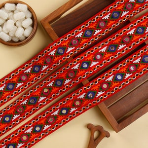 May include: Close-up of decorative red ribbon with embroidered details. The ribbon features a repeating pattern of colourful geometric shapes, small mirrors, and white stitching. The ribbon is likely used for embellishing crafts or clothing.