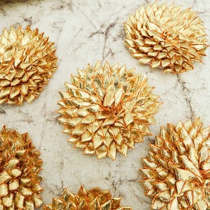 May include: Six gold-coloured, starburst-shaped embellishments are shown in a close-up view. Each has a textured, layered design, with a central point and radiating petals. They are arranged on a light, marbled surface, creating a decorative display.