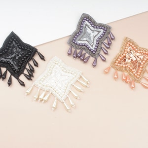 May include: Four square-shaped beaded embellishments with fringe. The embellishments are in black, white, light pink, and purple.