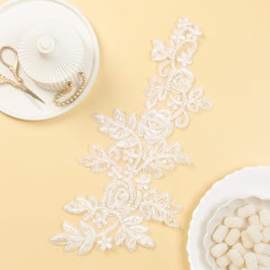 Puede incluir: Borde de encaje blanco con un diseño floral. El encaje presenta flores y hojas delicadas, creando un look romántico y elegante.