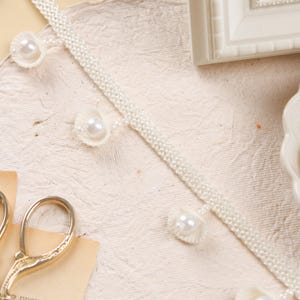 以下が含まれることがあります： アイボリー色の装飾トリム。織り模様に小さな丸いパールと貝殻型のアクセントが施されています。トリムは、テクスチャ加工されたオフホワイトの表面に表示されており、手芸品を示唆しています。繊細でヴィンテージな美学を醸し出しています。