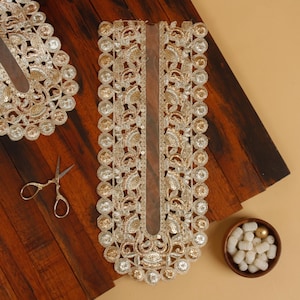 Puede incluir: Primer plano de un adorno de encaje dorado con intrincados detalles florales y lentejuelas. También se ven unas tijeras doradas pequeñas y un cuenco de madera pequeño lleno de cuentas blancas. El adorno es probablemente para embellecer prendas o manualidades.