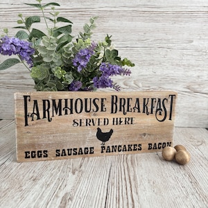 Peut inclure: Une enseigne en bois rustique avec le texte "Farmhouse Breakfast Served Here" et une silhouette de poulet. Sous le texte se trouvent les mots "Eggs Sausage Pancakes Bacon".