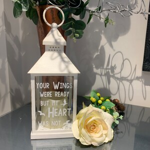 May include: White lantern with a glass window and a white candle inside. The lantern has the text "Your wings were ready but my heart was not" printed on it. A white rose is in the foreground.