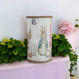 Op de afbeelding: Een witte cilindrische lampenkap met een jute rand heeft een gedrukt beeld van Peter Rabbit uit het kinderboek van Beatrix Potter. De afbeelding toont Peter Rabbit die in een tuin staat met bloemen en een vlinder.
