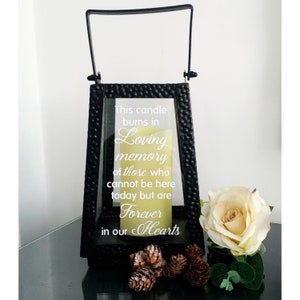 Puede incluir: Linterna de metal negro con un panel de vidrio que presenta el texto "This candle burns in loving memory of those who cannot be here today but are forever in our hearts". Una vela amarilla está dentro de la linterna. Una sola rosa blanca y piñas están en primer plano.