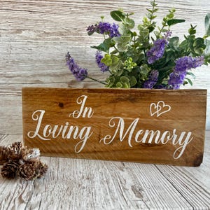 Puede incluir: Un letrero de madera con el texto "In Loving Memory" en letras blancas cursivas. Dos corazones blancos están por encima del texto. El letrero está sobre una superficie de madera blanca con un ramo de flores moradas detrás.