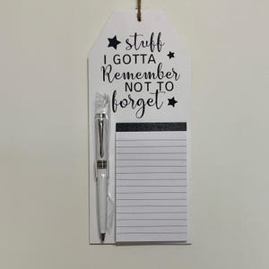 May include: White wooden sign with black text that reads "stuff i gotta remember not to forget" with a black star on each side of the text. A white pen and a notepad are attached to the sign.