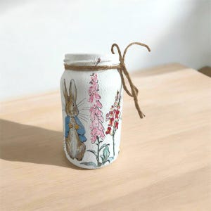Può includere: Un barattolo di vetro dipinto di bianco con un fiocco di spago marrone. Il barattolo presenta un'illustrazione dipinta a mano di Peter Rabbit dal libro per bambini di Beatrix Potter, circondato da fiori rosa e rossi.