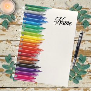 May include: A white notebook with a spiral binding on the left side. The notebook has a rainbow of coloured markers on the left side and the word "Name" written in cursive on the right side. A black pen is lying on a wooden surface next to the notebook.
