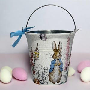 May include: A white metal bucket with a blue ribbon and a Peter Rabbit design. The bucket is decorated with flowers and butterflies. There are four pastel-colored eggs in the foreground.
