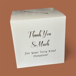 May include: A white donation box with a slot on top. The box has the words "Thank You So Much For Your Very Kind Donation!" printed in black script. The box is square and sits on a brown surface.