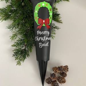Może przedstawiać: Czarny plastikowy wazon na kwiaty bożonarodzeniowe z zielono-brązowym wzorem wieńca i czerwoną kokardą. Tekst "Merry Christmas Dad" jest napisany białą czcionką. Wazon ma kształt stożka ze spiczastą podstawą.