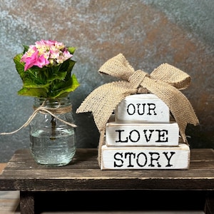 May include: A decorative arrangement on a wooden surface. A mason jar with pink and green flowers is tied with twine. Beside it, three white wooden blocks spell "OUR LOVE STORY", topped with a burlap bow. The arrangement is rustic in style.