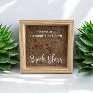 May include: A wooden frame with a glass front, containing a printed message that reads "In case of Dementia or Death Break Glass". The background is brown paper with a floral pattern.