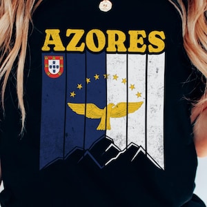May include: Black t-shirt featuring the word "AZORES" in yellow block letters above a graphic of the Azores flag. The flag design includes the Portuguese flag, a blue and white striped section with yellow stars, and a golden bird emblem.