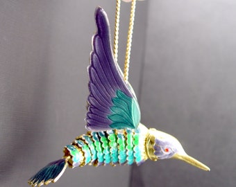 May include: A gold hummingbird ornament with purple and green wings. The hummingbird is hanging from a gold chain.