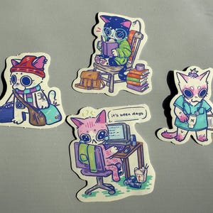 May include: Three sticker designs featuring cartoon cats. The first sticker shows a white cat with pink ears and a red hat carrying a brown bag. The second sticker shows a black cat with green eyes wearing a green shirt and reading a book. The third sticker shows a white cat with pink ears wearing a blue shirt and holding a cup.