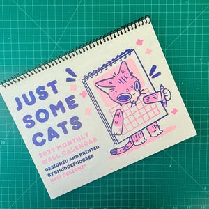 May include: A 2027 monthly wall calendar with a black spiral binding. The cover features the text "JUST SOME CATS 2027 MONTHLY WALL CALENDAR DESIGNED AND PRINTED BY SMUDGEPUDGEEE NEW DESIGNS!" and a pink and purple illustration of a cat holding a pencil.