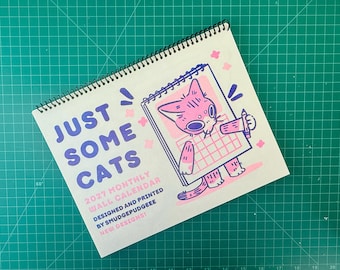 May include: A 2027 monthly wall calendar with a black spiral binding. The cover features the text "JUST SOME CATS 2027 MONTHLY WALL CALENDAR DESIGNED AND PRINTED BY SMUDGEPUDGEEE NEW DESIGNS!" and a pink and purple illustration of a cat holding a pencil.