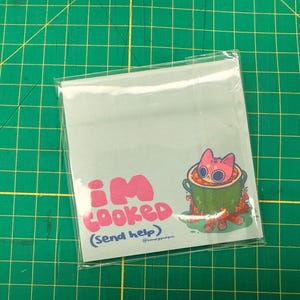 May include: A light blue square notepad with the text "I'M COOKED (Send help)" in pink. A cartoon cat with pink fur is in a green pot over a fire. The notepad is in a clear plastic sleeve.