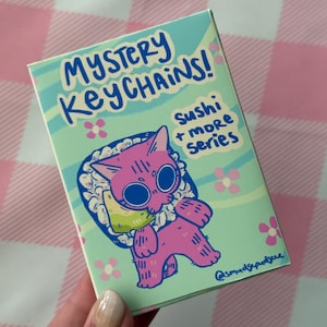 May include: A rectangular box featuring a pink cat designed as sushi, with large eyes and a cute expression. The box has the text "MYSTERY KEYCHAINS!" and "Sushi + more Series" in a playful font. The background is light green with flower illustrations.