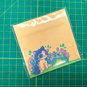 May include: A square, light orange art print in a clear plastic sleeve. The print features a cartoon cat with large glasses sitting in a field of pink tulips and green foliage. The background is a grid pattern.