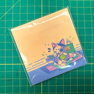 May include: A square notepad in a clear plastic sleeve. The notepad features a cartoon cat knitting with a yarn bowl and a fireplace. The background is light orange and blue.