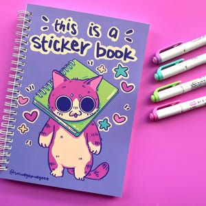 May include: A spiral-bound sticker book with a purple cover featuring a cartoon cat holding a green notebook. The text "this is a sticker book" is above the cat. Four white highlighters with different colored caps are on the right.