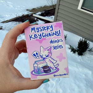 May include: A pink rectangular box with the text "MYSTERY KEYCHAINS! drinks series" in blue. The box features a cartoon cat with a bandage, a milk carton, and an Oreo cookie. The background has pink floral shapes.