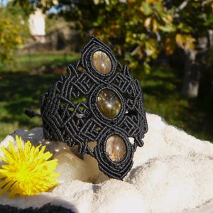 Puede incluir: Pulsera de macramé negra con tres piedras ovaladas de color ámbar. La pulsera presenta un diseño geométrico y se exhibe sobre una superficie blanca con una flor amarilla en primer plano. El fondo está borroso.