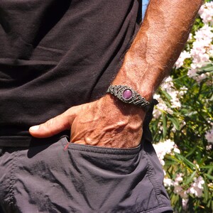 Bracciale a manetta intrecciato a mano in Macramè filato color grigio-verde con pietra naturale Rubino Stella indossato da uomo con mano in tasca sullo sfondo fiori di Oleandro.