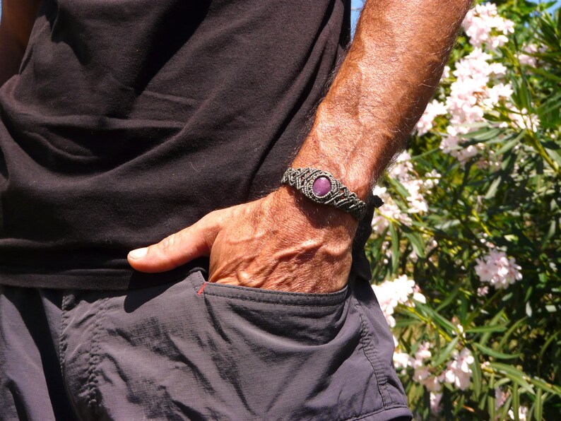Bracciale a manetta intrecciato a mano in Macramè filato color grigio-verde con pietra naturale Rubino Stella indossato da uomo con mano in tasca sullo sfondo fiori di Oleandro.