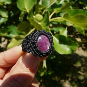 Primo piano laterale destro di anello nero, con al centro grande pietra preziosa naturale viola Rubino Stella, tenuto in mano su sfondo verde natura, alla luce del sole.