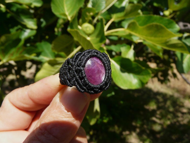 Primo piano laterale destro di anello nero, con al centro grande pietra preziosa naturale viola Rubino Stella, tenuto in mano su sfondo verde natura, alla luce del sole.