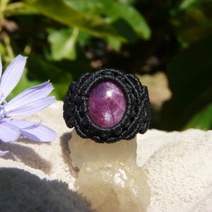Primo piano di anello nero, con al centro grande pietra preziosa naturale viola Rubino Stella, poggiato sopra minerale grezzo bianco con fiore azzurro, su sfondo naturale verde alla luce del sole.