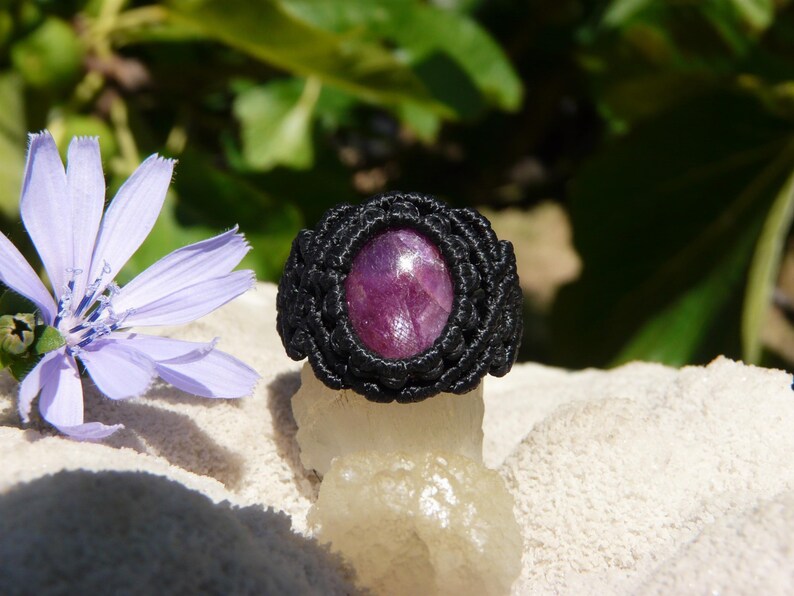 Primo piano di anello nero, con al centro grande pietra preziosa naturale viola Rubino Stella, poggiato sopra minerale grezzo bianco con fiore azzurro, su sfondo naturale verde alla luce del sole.
