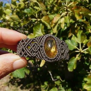Peut inclure: Un bracelet en macramé avec une pierre d'ambre ovale au centre. Le bracelet est fait de cordon marron foncé, présentant un motif tissé. La pierre d'ambre a une teinte dorée, sur un fond de feuillage vert flou.