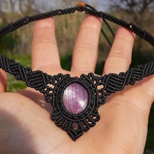 Può includere: Una collana girocollo in macramè nero con una pietra centrale ovale di colore viola chiaro. La collana è tenuta in una mano, evidenziando l'intricato dettaglio dei nodi e le variazioni di colore della pietra.