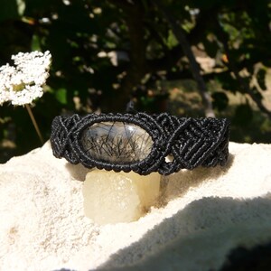 Puede incluir: Pulsera de macramé negro con una piedra ovalada transparente con inclusiones negras. La pulsera se exhibe sobre una superficie blanca texturizada, con un fondo borroso de vegetación y flores blancas.