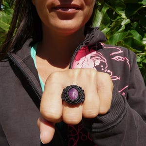 Primo piano di anello nero, con al centro grande pietra preziosa naturale viola Rubino Stella, indossato con mano chiusa da donna con felpa nera su sfondo verde natura, alla luce del sole.