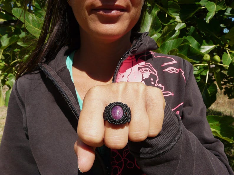 Primo piano di anello nero, con al centro grande pietra preziosa naturale viola Rubino Stella, indossato con mano chiusa da donna con felpa nera su sfondo verde natura, alla luce del sole.