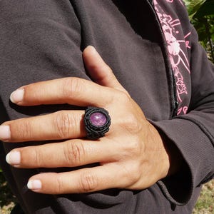 Primo pieno di anello nero con pietra preziosa ovale viola Rubino, indossato da donna con felpa nera, su sfondo verde alla luce del sole.